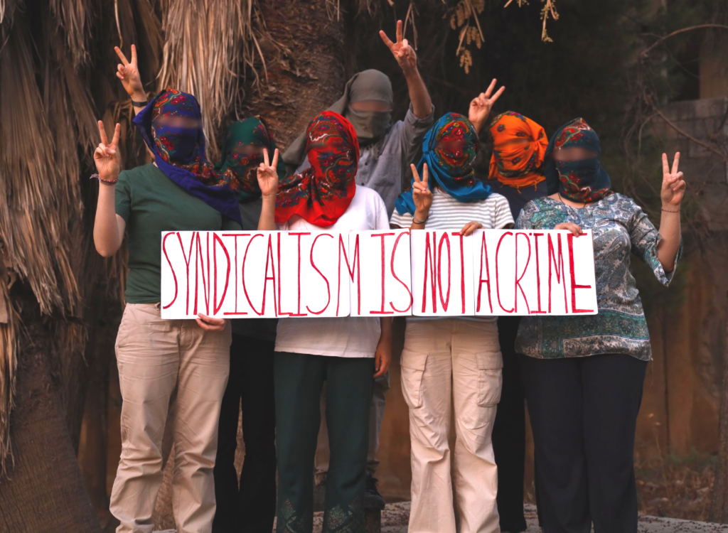 Six friends from the Internationalist Commune of Rojava standing together holding a banner with the words "Solidarity is not a crime!" on it. Thez stand in front of a palm tree.